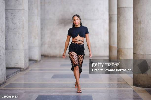 Ellie Delphine wears gold earrings, a black cropped / heart neck / laces waist t-shirt, a black pearls mesh / checkered tube knees skirt, black...