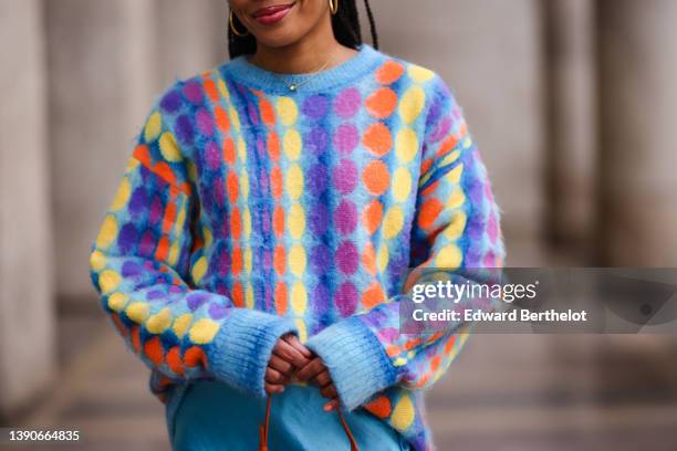 Ellie Delphine wears gold earrings, a gold chain pendant necklace, a pale blue with yellow / orange / purple circle print pattern wool pullover, a...