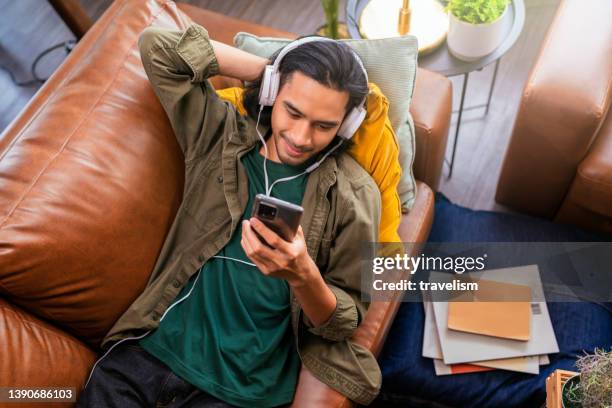 hombre asiático con auriculares escuchando la música y gesticulando mientras está sentado en su sofá.clam y tranquilo en casa alegre joven asiático con auriculares y listas de reproducción de teléfonos inteligentes elige reproducir canciones en el t - headphones asian fotografías e imágenes de stock