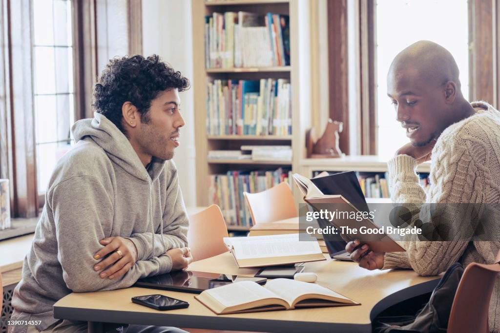 Male friends sitting at table talking to each other in library