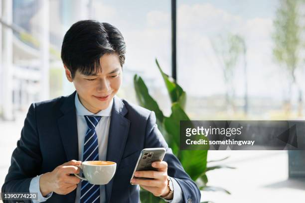 un joven empresario asiático mira su celular en una cafetería - toma mediana fotografías e imágenes de stock