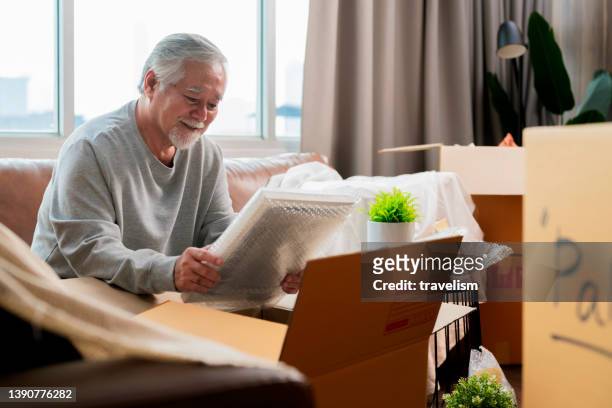 horizontale mittlere aufnahme eines erwachsenen älteren asiatischen mannes männlicher weißer bart, der auf dem boden sitzt und sich auf den umzug in ein neues haus vorbereitet, dinge in kisten packt und sie im wohnzimmer klebt, konzept für umzugsideen - entrümpeln stock-fotos und bilder