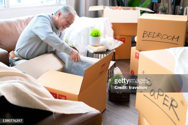 horizontal medium shot of adult elder senior asian man male white beard sitting on floor preparing for moving to new house packing things into boxes and taping them in  living room,home moving ideas concept - senior moving house stock pictures, royalty-free photos & images