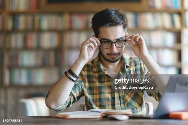 junger mann mit brille in der bibliothek, der auf einen laptop schaut - astigmatism stock-fotos und bilder