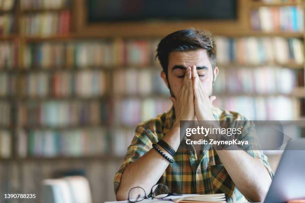 müder junge mann reibt sich die augen in der bibliothek - astigmatism stock-fotos und bilder