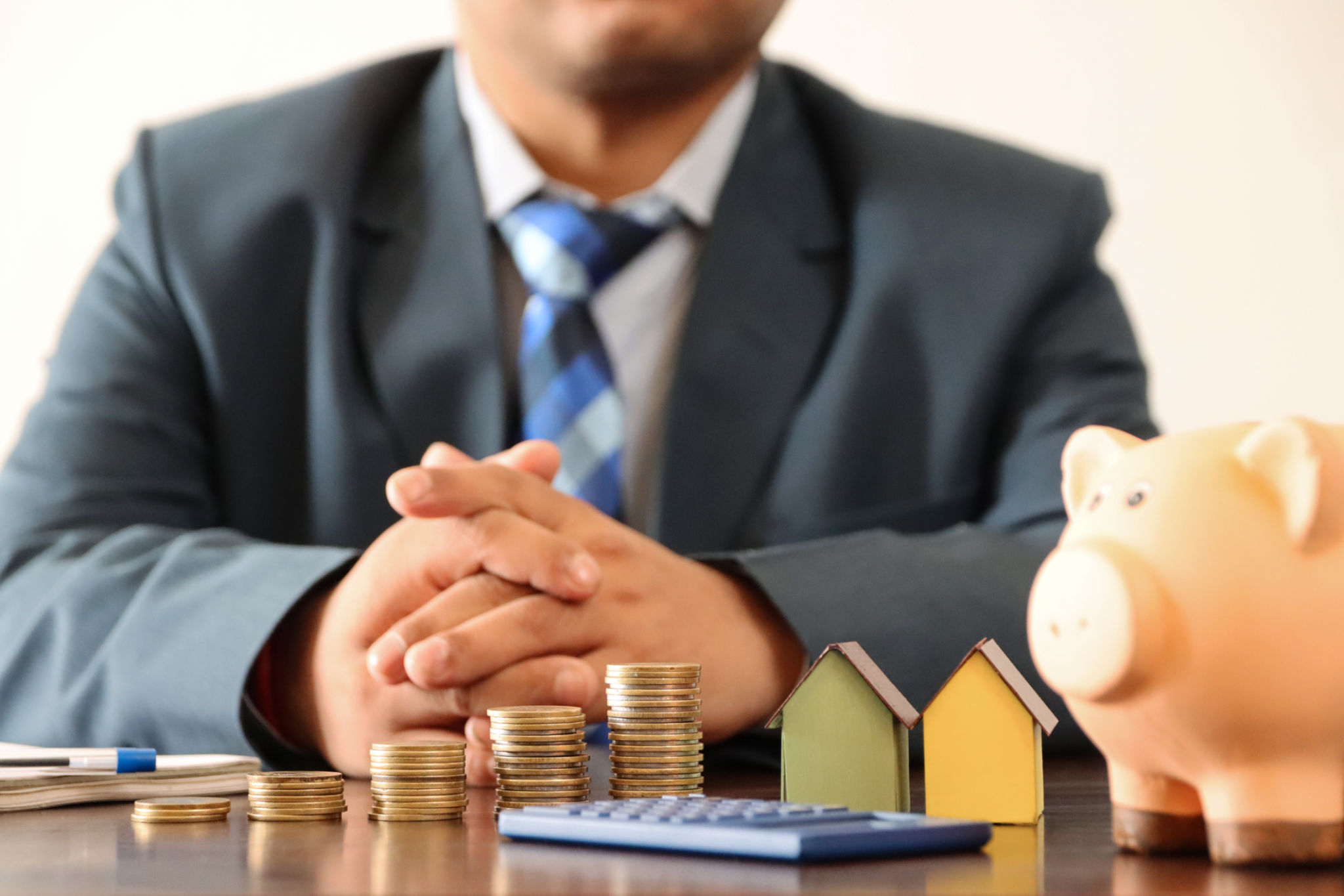 Close-up image of unrecognisable business man wearing smart, blue suit with white, button down collared shirt and blue checked tie sat behind desk, stacked coins, calculator, piggy bank, cardboard houses, real estate and finance concept Close-up image of unrecognisable business man wearing smart, blue suit with white, button down collared shirt and blue checked tie sat behind desk, stacked coins, calculator, piggy bank, cardboard houses, real estate and finance concept