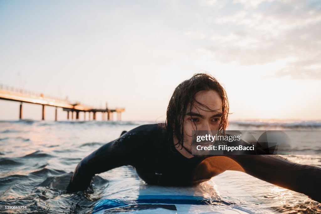 A man is swimming with his surfboard in the sea at sunset