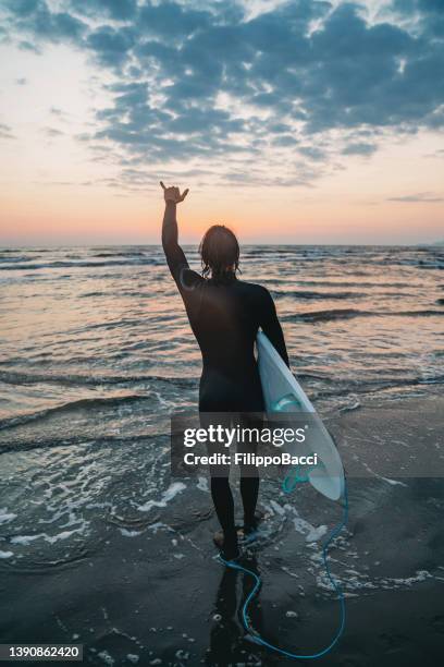 rear view of a man holding his surfboard after sunset and making the shaka sign to the sea - shaka stock pictures, royalty-free photos & images