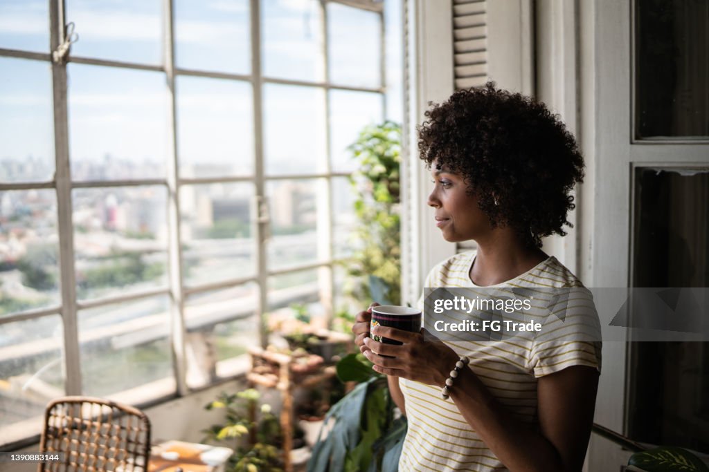 Kontemplative junge Frau, die einen Becher hält und zu Hause durch das Fenster schaut