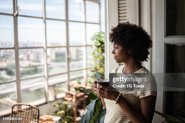 giovane donna contemplativa che tiene una tazza e guarda attraverso la finestra di casa - guardare fuori dalla finestra foto e immagini stock