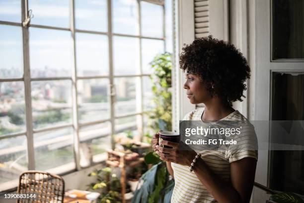 contemplative young woman holding a mug and looking through the window at home - coffe house live stock pictures, royalty-free photos & images
