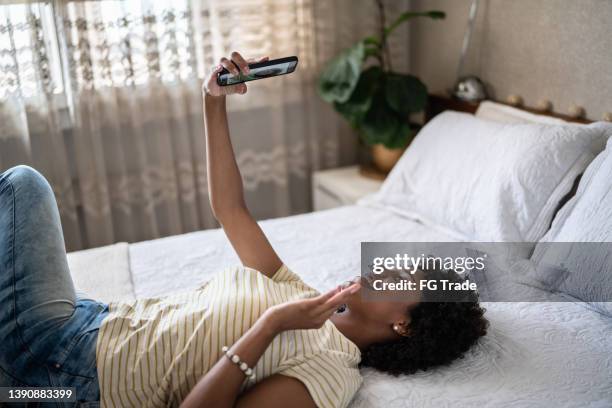 young woman on a video call on the mobile phone while lying down in the bed at home - lange afstandsrelatie stockfoto's en -beelden