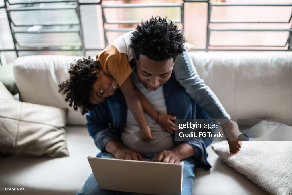 Father using the laptop trying to work while son is on his back at home