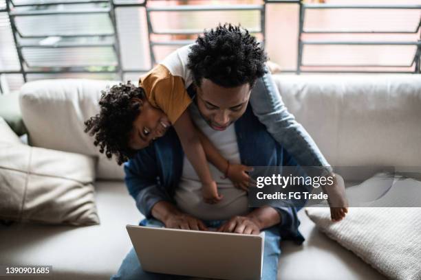 father using the laptop trying to work while son is on his back at home - social projects address needs of struggling families stockfoto's en -beelden