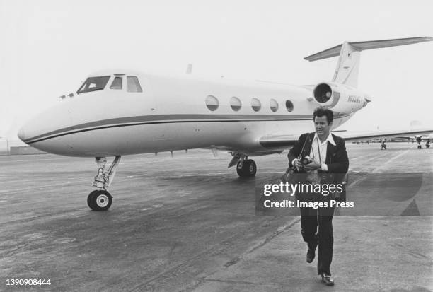 View of American photographer Ron Galella, with several cameras around his neck, as he walks on the tarmac at an unspecified airport, New York, 1970s.