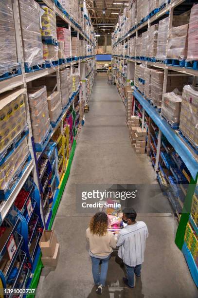 young couple pushing trolley down aisle of massive wholesaler - einkaufswagen voll stock-fotos und bilder