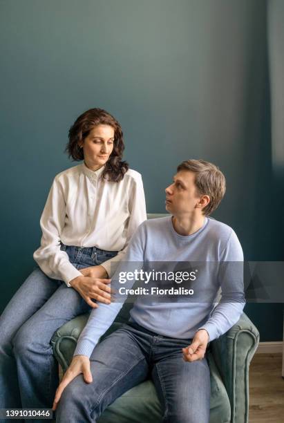 a man and a woman in a white shirt and blue jeans are sitting on an armchair against a wall. husband and wife, family full-length portrait - couple lifestyle jean stock pictures, royalty-free photos & images