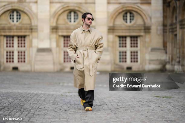 Alejandro Acero wears black sunglasses, a white t-shirt, a beige long belted trench coat, black large flared suit pants, yellow / orange print...