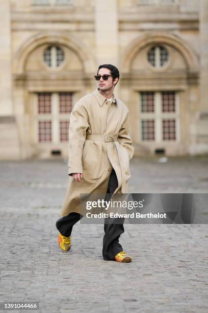 Alejandro Acero wears black sunglasses, a white t-shirt, a beige long belted trench coat, black large flared suit pants, yellow / orange print...