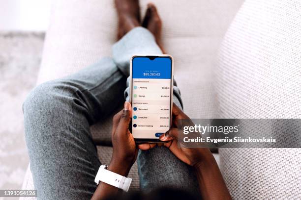 overhead view of young black woman managing online banking with smartphone sitting on the sofa at home. - cuenta de banco fotografías e imágenes de stock