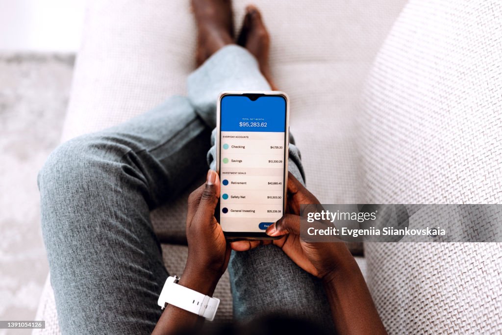 Overhead view of young Black woman managing online banking with smartphone sitting on the sofa at home.