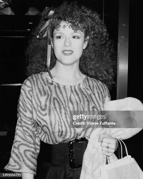 American actress and singer Bernadette Peters, carrying a jacket and a purse, wearing a ribbon in her hair, circa 1985.