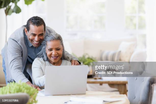 husband looks over wife's shoulder at grandchildren's photos on laptop - senior couple stockfoto's en -beelden