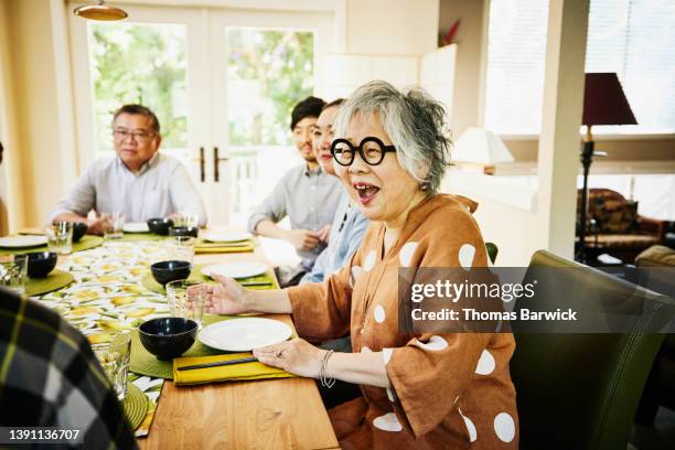 medium wide shot of smiling grandmother seated at dining room table for multigeneration family dinner - elderly asian foto e immagini stock