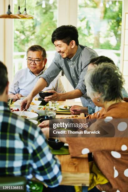 medium wide shot of smiling adult son severing himself food during multigenerational family dinner - medium group of people fotografías e imágenes de stock