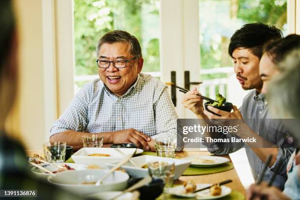 medium wide shot of laughing grandfather seating at head of dinning room table during multigenerational family dinner - elderly chinese man stock-fotos und bilder