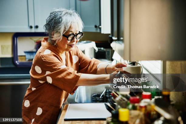 medium wide shot of senior woman cleaning up in kitchen after dinner with family - asian woman beauty shot photos et images de collection