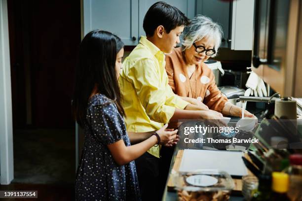 medium wide shot of grandchildren helping grandmother with dishes in kitchen after family dinner party - kitchen after party stock-fotos und bilder