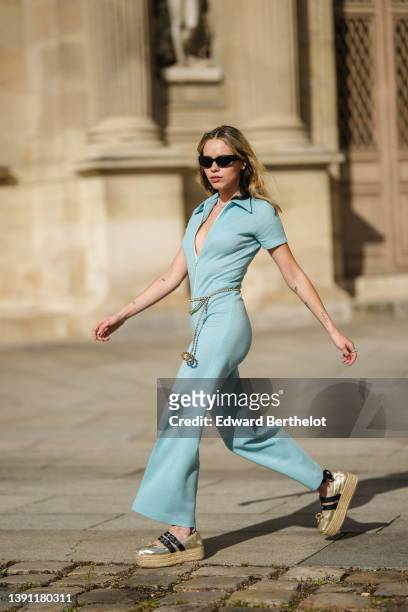 Almuneda Lapique wears black sunglasses, a silver rhinestones necklace, a pale blue ribbed short sleeves / white zipper / flared jumpsuit, a...