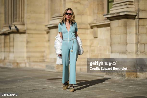 Almuneda Lapique wears black sunglasses, a silver rhinestones necklace, a pale blue ribbed short sleeves / white zipper / flared jumpsuit, a...