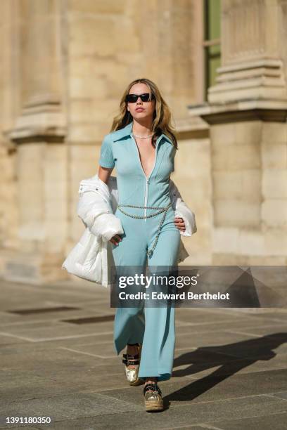 Almuneda Lapique wears black sunglasses, a silver rhinestones necklace, a pale blue ribbed short sleeves / white zipper / flared jumpsuit, a...