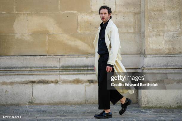 Lukas Osiecki wears a black shirt, a white latte long coat, black large suit pants, black shiny leather loafers from Prada, during a street style...