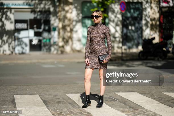 Emy Venturini wears black sunglasses, gold fringed chain pendant earrings, a brown / black leopard print pattern turtleneck tube short dress, a black...