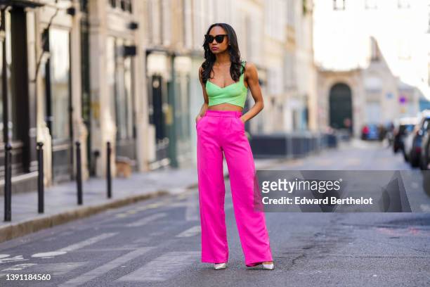 Emilie Joseph @in_fashionwetrust wears black sunglasses, silver rhinestones earrings, neon pink large suit pants, a pale green cross-strappy V-neck /...