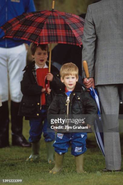British Royals Prince William holding a red tartan umbrella behind his brother Prince Harry, both wearing waxed jackets and wellington boots, attend...
