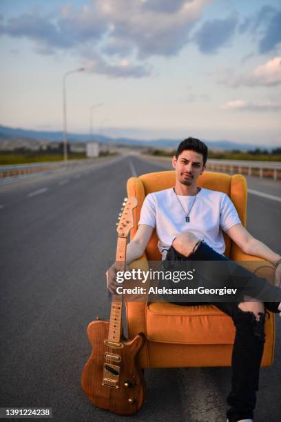 músico masculino relajado sentado en la silla y sosteniendo la guitarra en medio de la carretera - guitar case fotografías e imágenes de stock