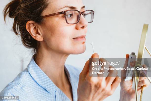 a woman looking her face in a mini mirror before applying an antiviral cream or ointment to treat herpes labialis on her lower lip. - cold sore stock-fotos und bilder