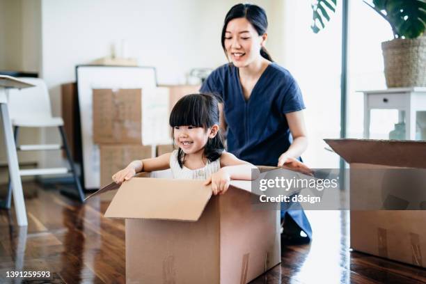 young asian family moving into a new apartment. mother is pushing the cardboard box while little daughter is sitting in the box having fun and smiling joyfully. home moving, migration, relocation concept - asian young family stock-fotos und bilder