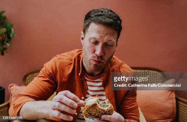 a man chews a large bite of a burger - enjoyment fotografías e imágenes de stock