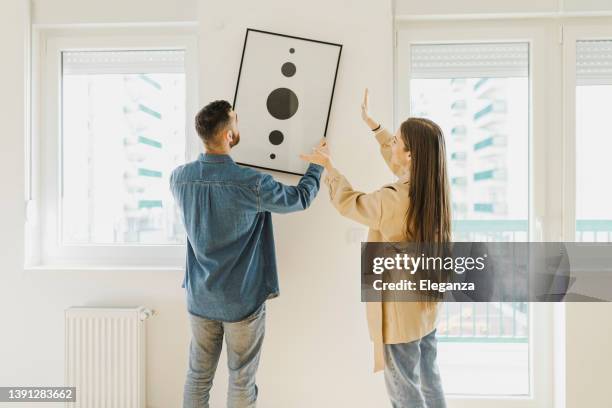 young couple arranging new flat, hanging up painting, new home and relocation concept. - draped stockfoto's en -beelden