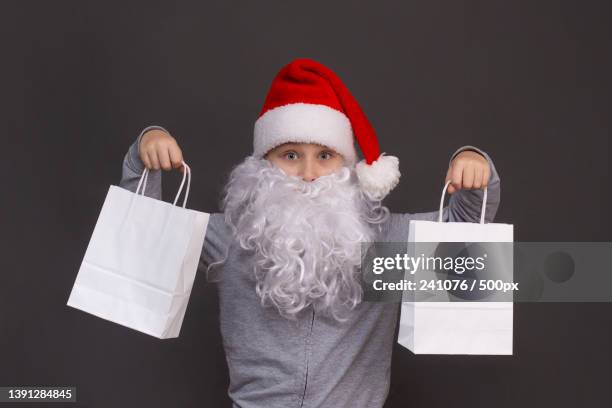 little santa boy is holding two white paper bags in his hands - santa hat and beard stock pictures, royalty-free photos & images