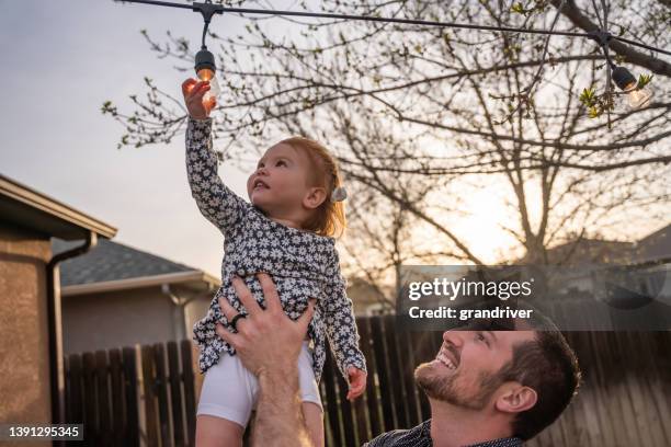 若い父親は、夕暮れ時に裏庭に装飾的に配置されたエジソン電球に触れると、2歳の娘を抱きしめる - 2 year old blonde girl father ストックフォトと画像