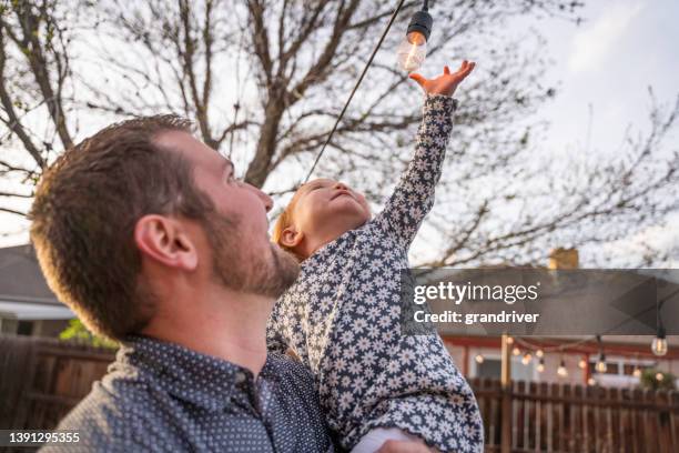若い父親は、夕暮れ時に裏庭に装飾的に配置されたエジソン電球に触れると、2歳の娘を抱きしめる - 2 year old blonde girl father ストックフォトと画像