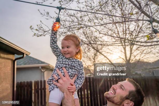 junger vater hält zweijährige tochter hoch, als sie eine edison-glühbirne berührt, die in der abenddämmerung dekorativ in einem hinterhof angeordnet ist - 2 year old blonde girl father stock-fotos und bilder