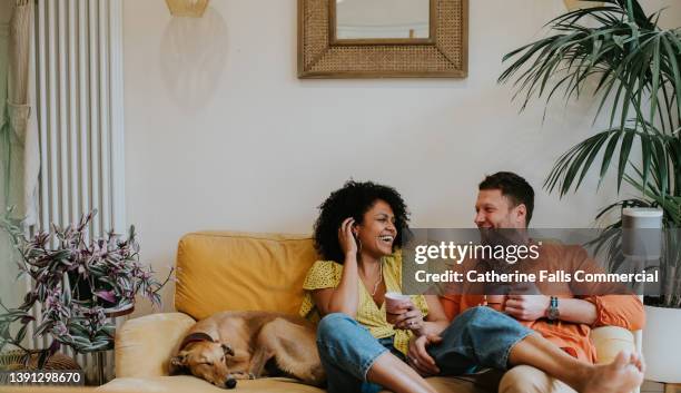 a beautiful, young, happy couple lounge together on a soft, luxurious yellow sofa in a modern living room. their dog naps beside them. - mühelosigkeit stock-fotos und bilder