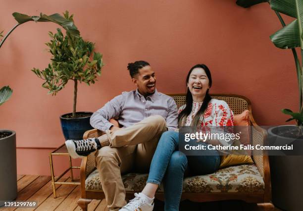 two friends / young couple sit on a sofa and laugh together - british asian couple stock pictures, royalty-free photos & images